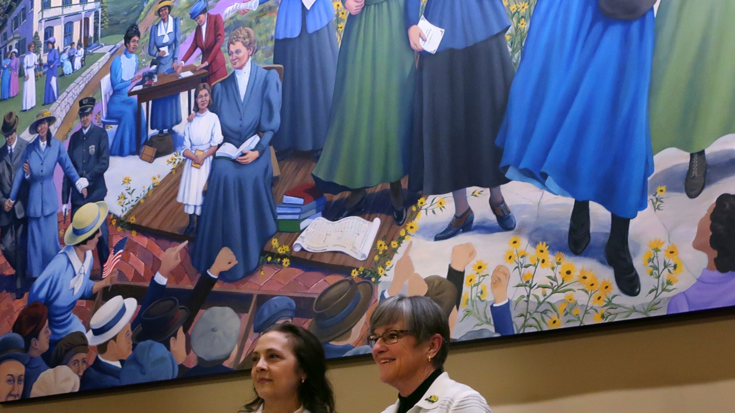 Artist Phyllis Garibay-Coon, left, and Kansas Gov. Laura Kelly, right, stand beneath Garibay-Coon's newly unveiled mural honoring Kansas women who campaigned for voting rights, Wednesday, Jan. 29, 2025, at the Statehouse in Topeka, Kan. (AP Photo/John Hanna)